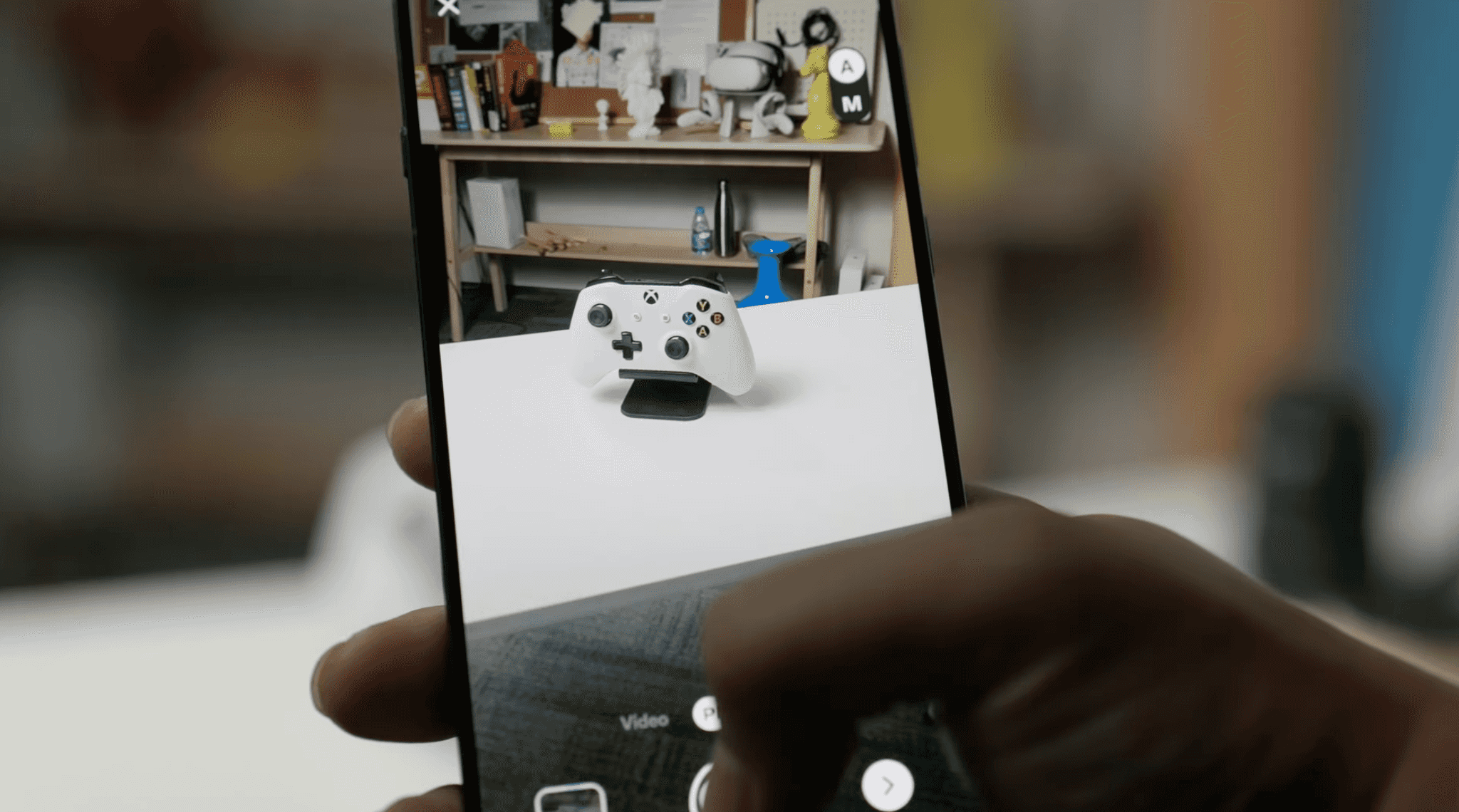 Hand holding a phone camera aimed at a small object on a table, illustrating photogrammetry capture with overlapping photos.